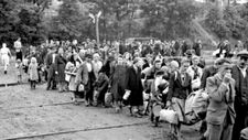 Sie stehen vor einer Wand mit einem 3 mal 4 ½ m großen Schwarzweißfoto. Darauf ist eine Menschenmenge abgebildet. Etwa hundert Personen, darunter Familien mit Kindern, gehen in einer Schlange hintereinander. Einige haben kleine Taschen dabei oder Kleidungsstücke in den Händen. Das Foto zeigt die Ankunft von Flüchtlingen im Lager Wipperfürth im Jahr 1947.