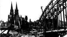 Vor Ihnen auf der Wand läuft eine etwa 3 ½ mal 3 m große Projektion mit verschiedenen Schwarzweißfotos. Sie zeigen das zerstörte Nordrhein-Westfalen nach dem Zweiten Weltkrieg, darunter die Stadt Köln. Im Hintergrund befindet sich der Kölner Dom, davor die völlig zerstörte Hohenzollernbrücke.